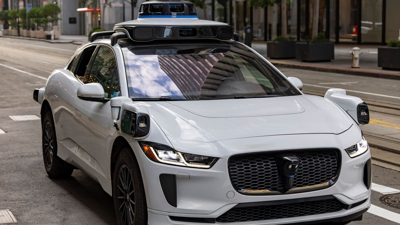 A white autonomous vehicle navigating a city street, reflecting urban architecture in daylight, representing 2026's robotaxi expansion